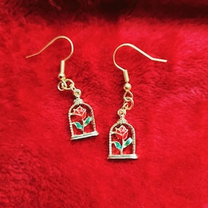 May include: Gold earrings with a red rose in a glass dome. The rose is surrounded by green leaves.