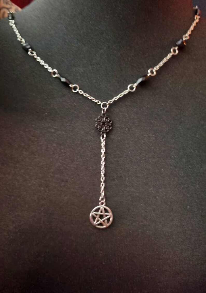 May include: A silver chain necklace with black beads and a silver pendant featuring a pentagram. The pendant hangs from a silver chain with a black filigree charm.