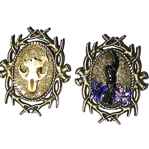 May include: Two oval-shaped pendants with bronze-colored frames. One pendant features a skull on a gold glitter background. The other pendant has a black object with purple flowers on a gold glitter background.
