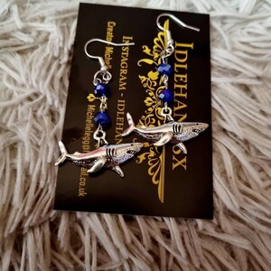 Pode incluir: Um par de brincos de tubar&atilde;o prateados com detalhes em cristal azul. Os brincos est&atilde;o pendurados em um cart&atilde;o preto com o texto "IDLEHANDS" e "INSTAGRAM - IDLEHANDS" nele.