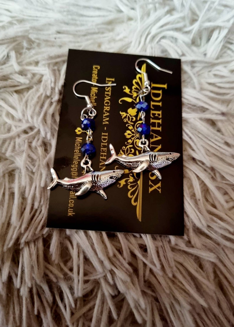Pode incluir: Um par de brincos de tubar&atilde;o prateados com detalhes em cristal azul. Os brincos est&atilde;o pendurados em um cart&atilde;o preto com o texto "IDLEHANDS" e "INSTAGRAM - IDLEHANDS" nele.
