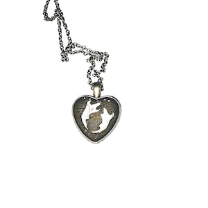 May include: A silver-toned necklace with a heart-shaped pendant. The pendant features a detailed design of two fish swimming in opposite directions, set against a textured background. The chain is delicate and the overall design is elegant.