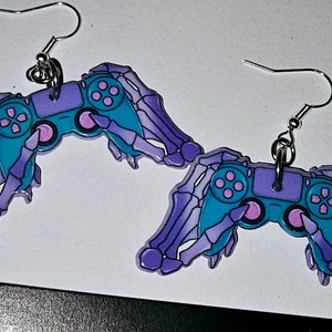 Könnte beinhalten: Ein Paar violette und blaue Skeletthand-Ohrringe mit einem Videospiel-Controller in jeder Hand. Die Controller sind blaugrün mit rosa Knöpfen.