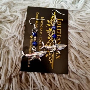 Pode incluir: Um par de brincos de tubar&atilde;o prateados com contas de cristal azul. Os brincos est&atilde;o pendurados em um cart&atilde;o preto com o texto "Idlehanx" e "Instagram - Idlehanx" impresso nele.