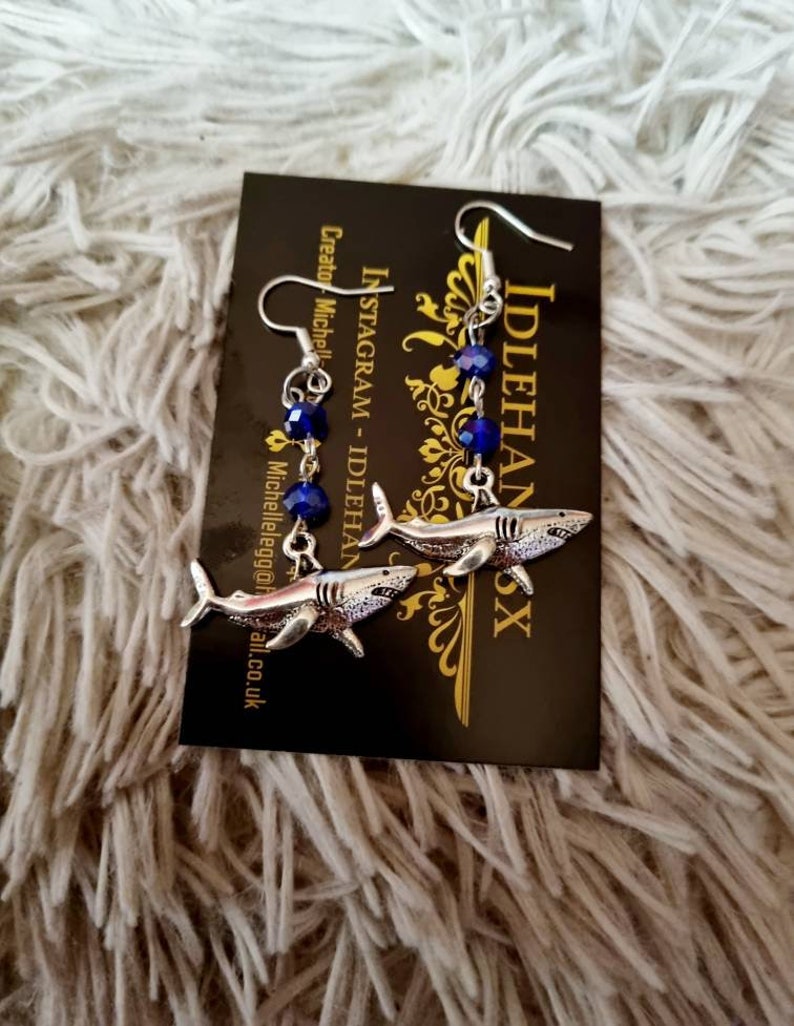 Pode incluir: Um par de brincos de tubar&atilde;o prateados com contas de cristal azul. Os brincos est&atilde;o pendurados em um cart&atilde;o preto com o texto "Idlehanx" e "Instagram - Idlehanx" impresso nele.
