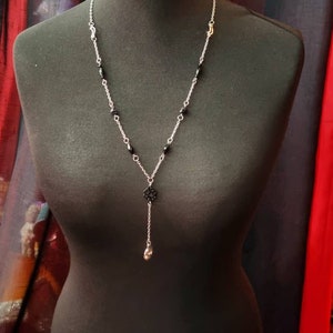 May include: A silver chain necklace with black beads and a silver pendant. The pendant is a small, intricate design with a skull at the bottom.
