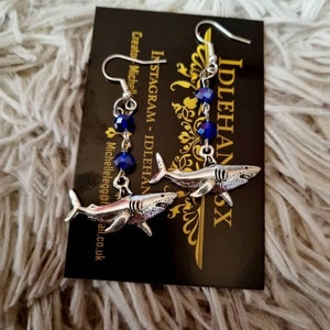 Pode incluir: Um par de brincos de tubar&atilde;o pendentes de prata com contas de cristal azul. Os brincos est&atilde;o em um cart&atilde;o preto com o texto "INSTAGRAM - IDLEHANDS" e "Criador: Michelle @idle.egg@gmail.co.uk".