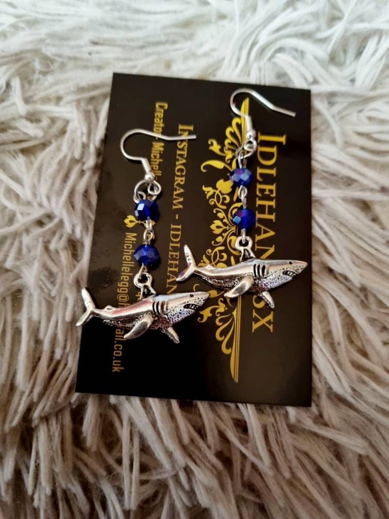 Pode incluir: Um par de brincos de tubar&atilde;o pendentes de prata com contas de cristal azul. Os brincos est&atilde;o em um cart&atilde;o preto com o texto "INSTAGRAM - IDLEHANDS" e "Criador: Michelle @idle.egg@gmail.co.uk".
