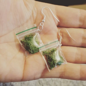 Peut inclure: Une paire de boucles d'oreilles fantaisie avec de petits sacs en plastique transparent remplis de matière verte, suspendus à des crochets d'oreille argentés. Les sacs ont un joint vert en haut.