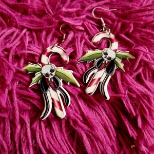 May include: A pair of silver dangle earrings with a candy cane design. The candy canes are red and white with a green holly leaf and a skull on each.