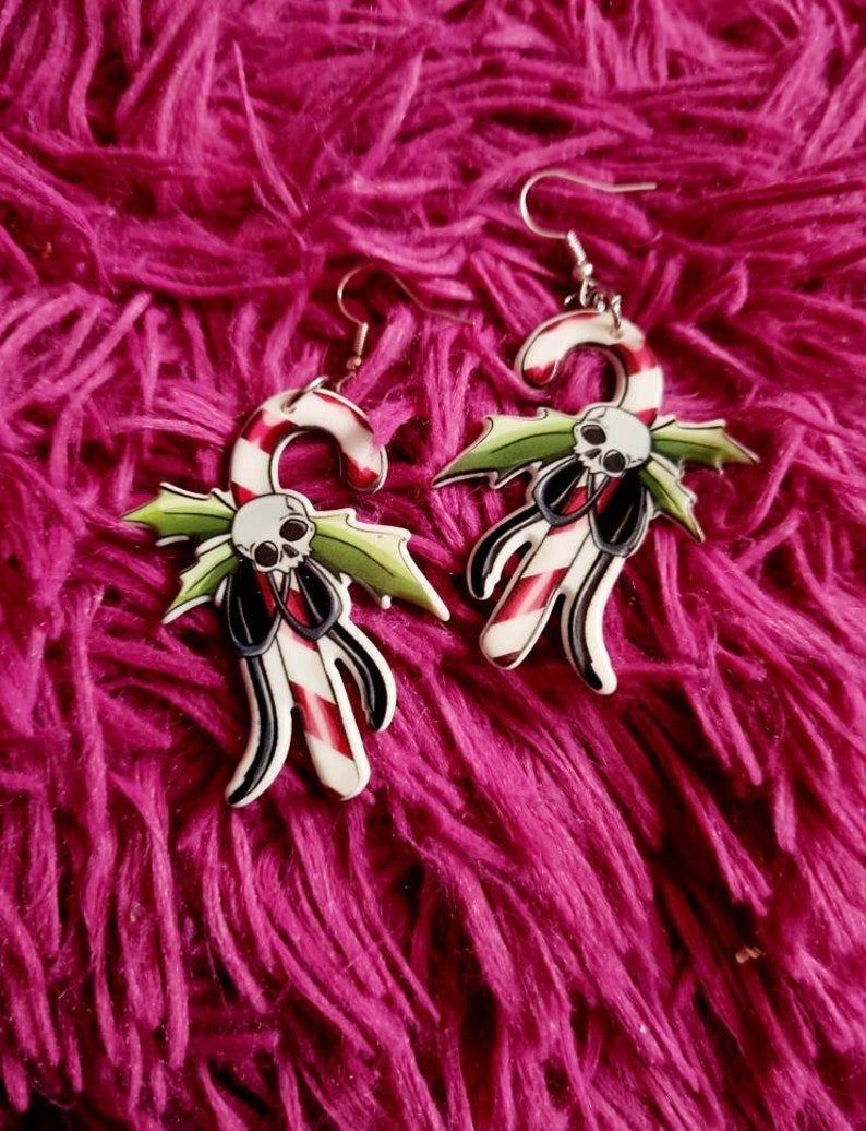 May include: A pair of silver dangle earrings with a candy cane design. The candy canes are red and white with a green holly leaf and a skull on each.