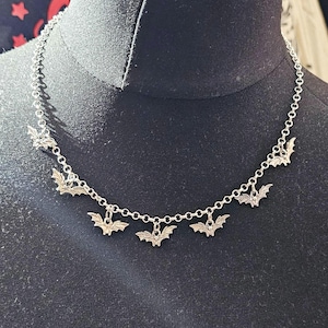 Könnte beinhalten: Silberfarbene Halskette mit Fledermaus-Anhängern. Die Halskette wird auf einer schwarzen Schaufensterpuppe präsentiert, die die zarte Kette und die detaillierten Fledermaus-Anhänger hervorhebt. Die Fledermäuse sind gleichmäßig verteilt.