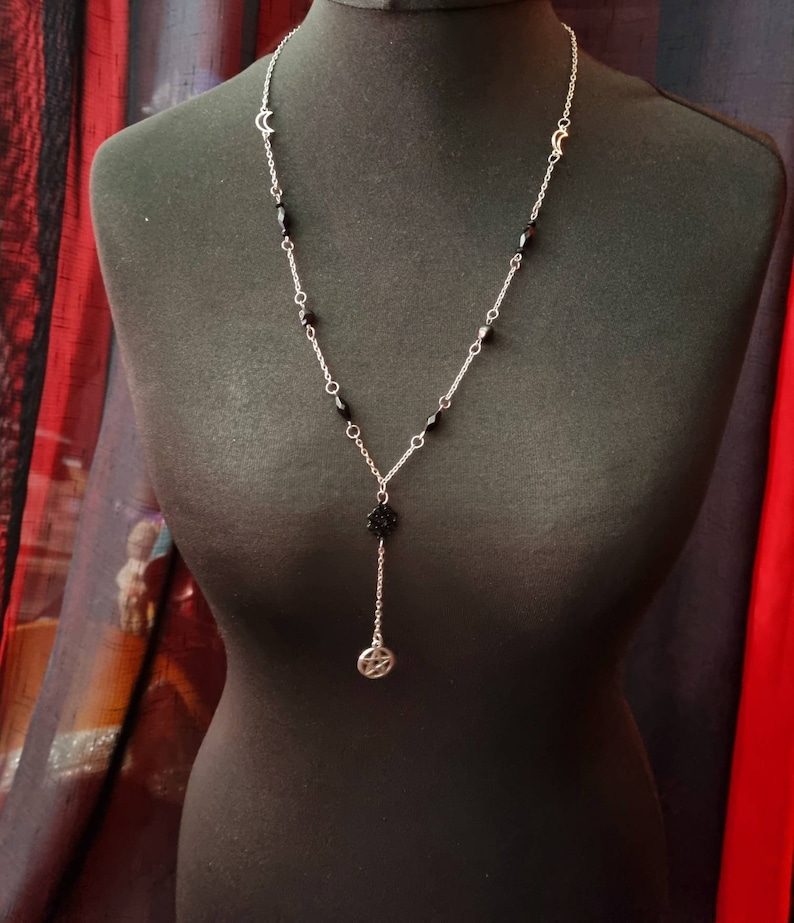May include: A silver chain necklace with black beads and a silver pentagram pendant. The necklace is displayed on a black mannequin.