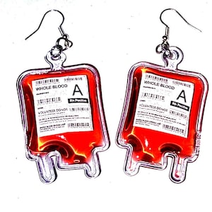 May include: A pair of novelty earrings shaped like blood bags. Each clear plastic bag is filled with red liquid and features a white label with the text "WHOLE BLOOD" and "A Rh Positive". The earrings have silver-colored ear wires.