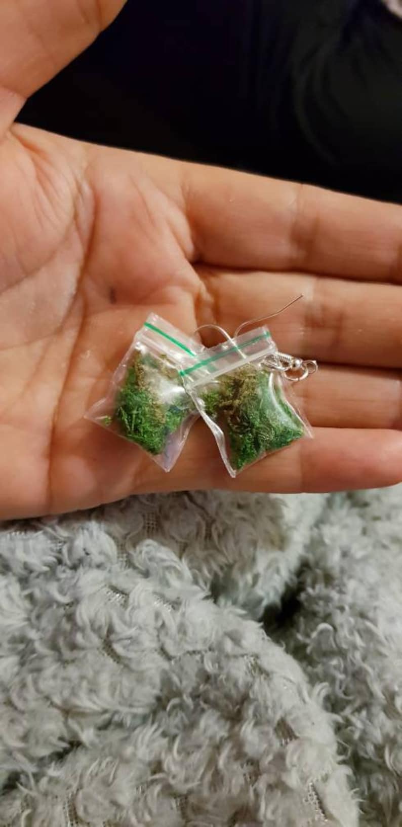 May include: A pair of novelty earrings featuring small clear plastic bags filled with green moss. The bags are sealed with a green stripe and attached to silver-colored earring hooks. The earrings are held in a hand.