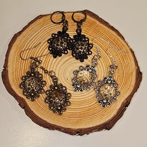 May include: Three pairs of earrings with a floral design and a round, textured, gold-toned centre piece. The earrings are displayed on a wooden surface.