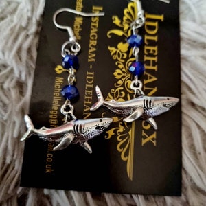 Pode incluir: Um par de brincos de tubar&atilde;o prateados com contas de cristal azul. Os brincos est&atilde;o pendurados em um cart&atilde;o preto com o texto "Instagram - Idlehands" e "Idlehands" nele.