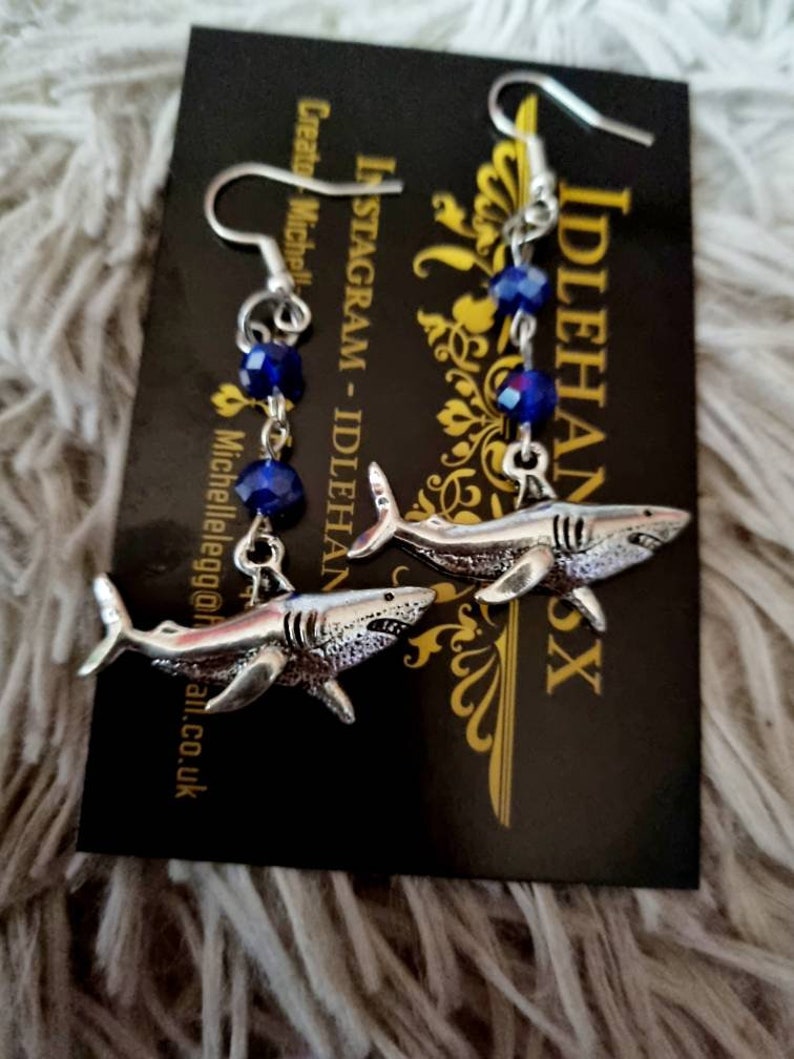 Pode incluir: Um par de brincos de tubar&atilde;o prateados com contas de cristal azul. Os brincos est&atilde;o pendurados em um cart&atilde;o preto com o texto "Instagram - Idlehands" e "Idlehands" nele.