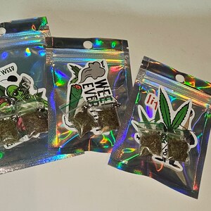 May include: Three small, clear plastic bags filled with dried plant material. Each bag is attached to a sticker with a cannabis-related design and text. The bags are arranged on a white surface.