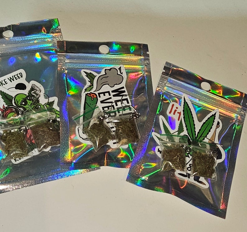 May include: Three small, clear plastic bags filled with dried plant material. Each bag is attached to a sticker with a cannabis-related design and text. The bags are arranged on a white surface.