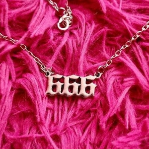 May include: A silver necklace with a pendant that says "666" in a gothic font. The necklace is on a pink fluffy surface.