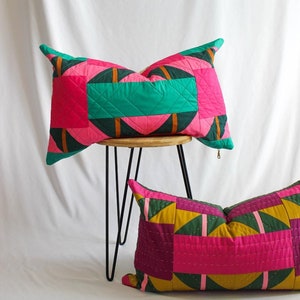 May include: Two decorative cushion covers featuring a geometric pattern in shades of pink, green and gold. The cushion covers are made from quilted fabric and have a zip closure.