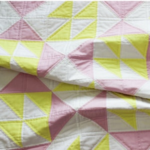 Puede incluir: Un patrón de colcha llamado "Pink Lemonade" con un diseño moderno. La colcha está hecha con tela rosa, amarilla y blanca. La colcha está doblada por la mitad y el patrón es visible en ambos lados. El texto "Skill Level | Easy" y "Shannon Fraser Designs" es visible en la parte inferior de la imagen.
