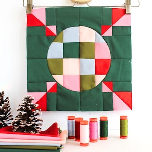 Pode incluir: Um bloco de colcha de tecido quadrado com um círculo central composto por uma grade multicolorida em tons de azul claro, verde oliva, rosa pálido e vermelho brilhante, sobre um fundo verde floresta escuro com detalhes em vermelho e rosa.