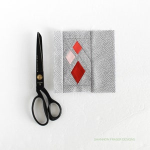 Może przedstawiać: Zbliżenie na blok kołdry z wzorem diamentowym w czerwonym, różowym i białym materiale. Blok jest zszyty na szarym materiale w białe kropki. Para czarnych nożyczek do tkanin leży na białym tle.