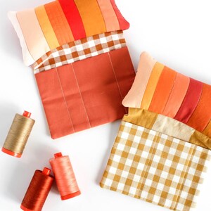 Puede incluir: Dos bolsas de tela hechas a mano con la parte superior de retazos coloridos y la parte inferior a cuadros o lisas. Las bolsas son en tonos naranja, rojo y melocotón. Bobinas de hilo en dorado, rojo y melocotón están esparcidas alrededor de las bolsas.