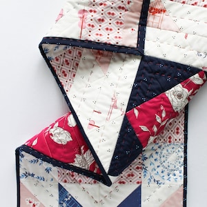 Puede incluir: Un corredor de colcha con un diseño de patchwork con telas rosas, rojas, azules y blancas. La colcha está bordeada con una cinta azul marino. La colcha está doblada por la mitad y los patrones de la tela son visibles en ambos lados. La colcha está hecha por Shannon Fraser Designs.