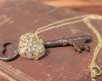 May include: A gold-colored heart-shaped pendant necklace with a floral design and small clear stones. The pendant is next to a vintage key and a keyhole-shaped object, all resting on a brown surface.