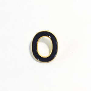 Può includere: Una spilla in metallo dorato a forma di lettera "O". La lettera è riempita di smalto blu navy scuro. La spilla è posta su uno sfondo bianco.
