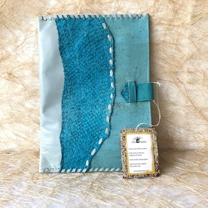 Puede incluir: Una carpeta de corcho azul y blanca con una correa de cuero y una etiqueta que dice "The Tree Garden, Porto de Pescas, Portugal,  Handmade from antique pieces,  Natural cork from Portugal,  The tree garden".