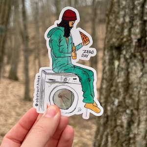 May include: A sticker featuring a cartoon illustration of a person wearing a red beanie and green jacket sitting on a white washing machine. The person is holding a slice of pizza and a can of beer. The sticker has the text "Zero Day" and "@abstract.hikes" on it.
