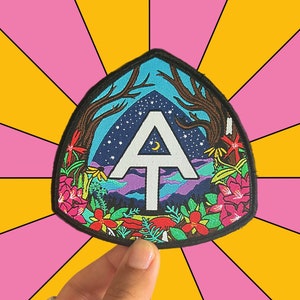 May include: A black and white embroidered patch with a white letter "A" with a crescent moon above it. The patch is surrounded by a colorful floral design with a blue sky and mountains in the background.