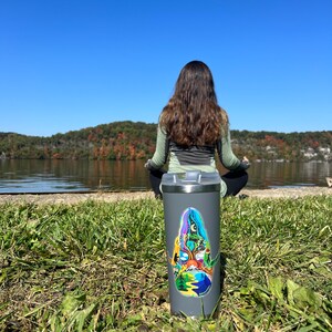 May include: A gray stainless steel travel mug with a colorful sticker featuring a nature scene. The sticker depicts a tree with a mountain range, a river, and a sun in the sky. The mug is sitting on a bed of green grass.