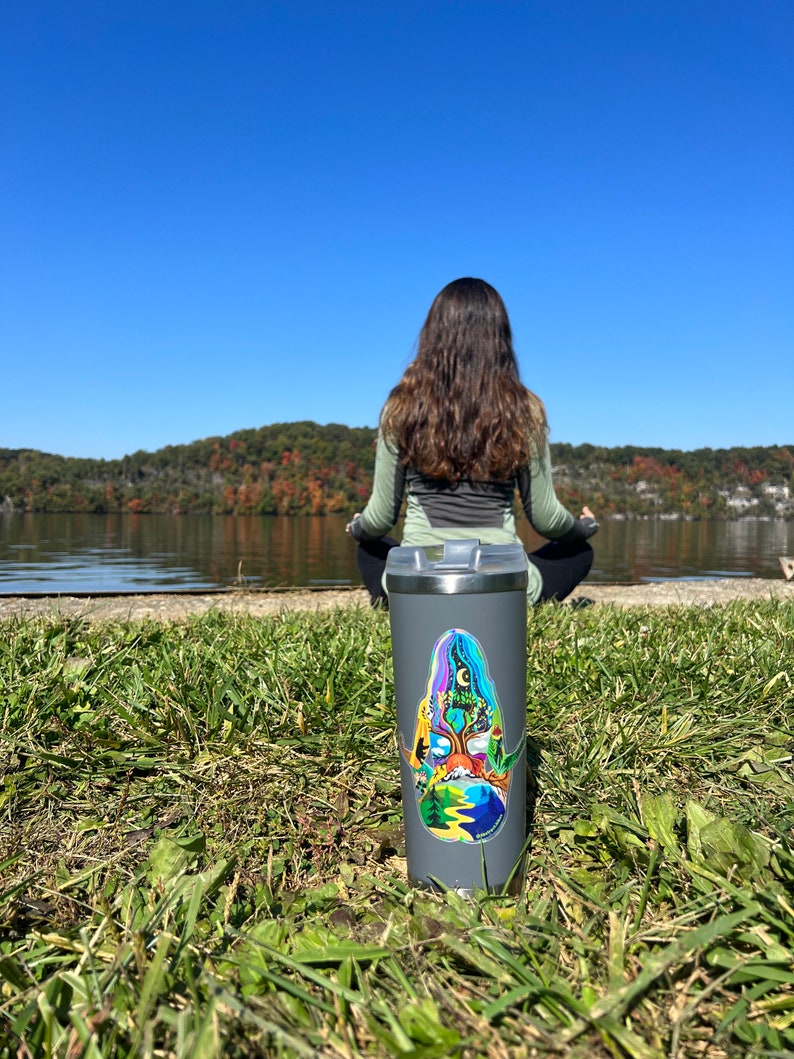 May include: A gray stainless steel travel mug with a colorful sticker featuring a nature scene. The sticker depicts a tree with a mountain range, a river, and a sun in the sky. The mug is sitting on a bed of green grass.