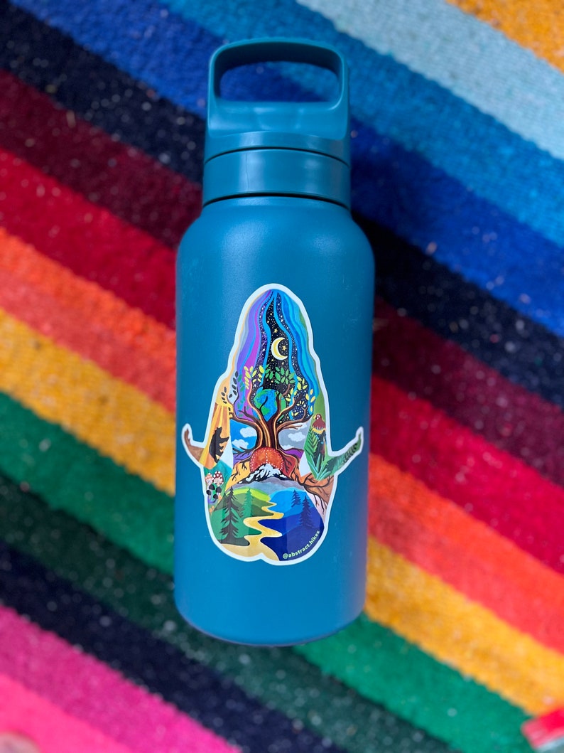 Puede incluir: Una botella de agua azul verdosa con una pegatina colorida de un &aacute;rbol con una luna y estrellas en el fondo. La pegatina tiene el texto "@abbyfrancesart" en la parte inferior.