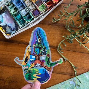 May include: A holographic sticker featuring a colorful illustration of a person meditating with a tree growing from their head. The tree is surrounded by nature scenes, including mountains, a river, and a moon. The sticker has the text "@abstract.hikes" at the bottom.