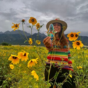 Puede incluir: Una persona con un su&eacute;ter a rayas de colores y un sombrero de ala ancha se encuentra en un campo de girasoles amarillos. Est&aacute; sosteniendo una pegatina con un dise&ntilde;o colorido.
