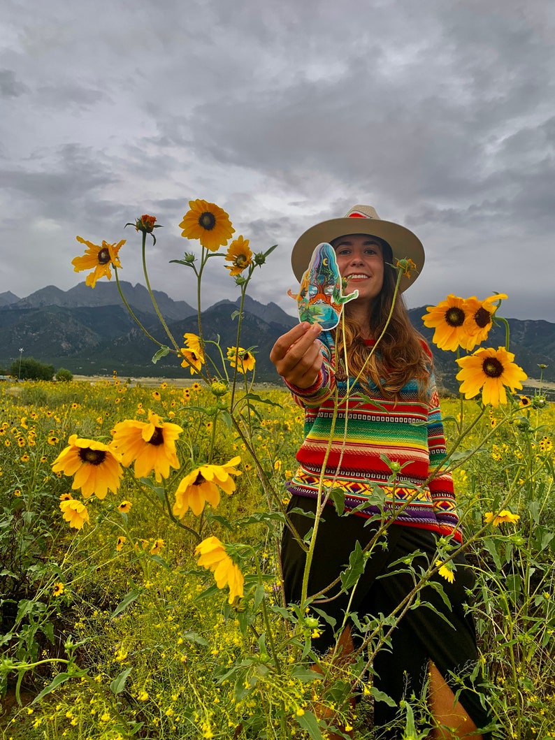 Puede incluir: Una persona con un su&eacute;ter a rayas de colores y un sombrero de ala ancha se encuentra en un campo de girasoles amarillos. Est&aacute; sosteniendo una pegatina con un dise&ntilde;o colorido.