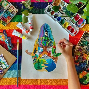 Puede incluir: Una persona pinta una ilustraci&oacute;n colorida de un &aacute;rbol con una figura humana en un lienzo blanco. La ilustraci&oacute;n est&aacute; rodeada de varios materiales de arte, incluyendo pinceles, paletas de acuarela y tubos de pintura.