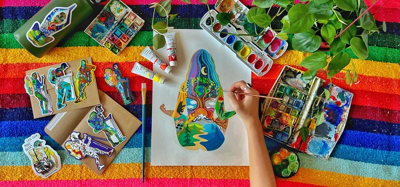Puede incluir: Una persona pinta una ilustraci&oacute;n colorida de un &aacute;rbol con una figura humana en un lienzo blanco. La ilustraci&oacute;n est&aacute; rodeada de varios materiales de arte, incluyendo pinceles, paletas de acuarela y tubos de pintura.
