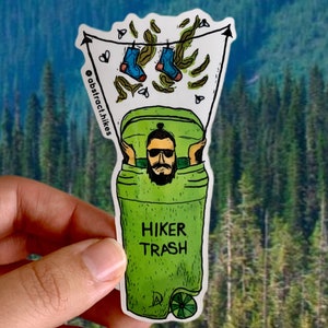 Puede incluir: Un contenedor de basura verde con las palabras "Hiker Trash" impresas en él. Un excursionista de dibujos animados está dentro del contenedor de basura, con gafas de sol y sosteniendo un par de calcetines azules. El contenedor de basura está sobre un fondo blanco con hojas verdes y calcetines azules.