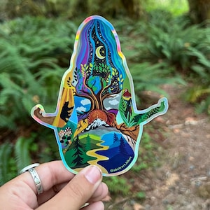 May include: A holographic sticker featuring a colorful illustration of a tree with a mountain, a river, and a bear. The sticker is shaped like a person in a yoga pose. The text "@abstract.hikes" is at the bottom of the sticker.