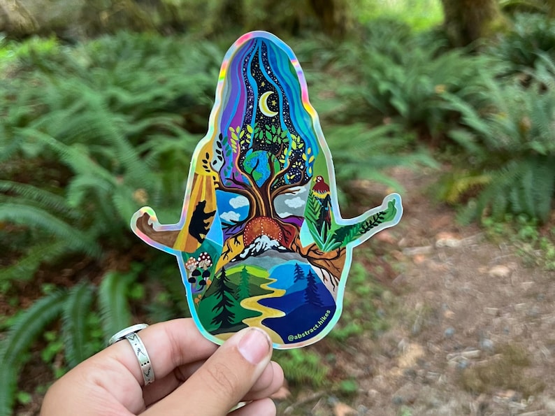 May include: A holographic sticker featuring a colorful illustration of a tree with a mountain, a river, and a bear. The sticker is shaped like a person in a yoga pose. The text "@abstract.hikes" is at the bottom of the sticker.
