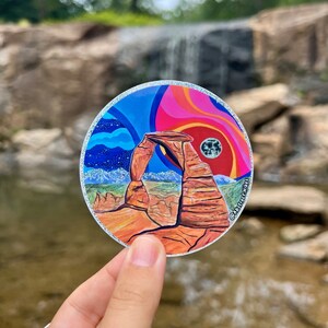 Könnte beinhalten: Ein runder Aufkleber mit einer farbenfrohen Illustration des Delicate Arch im Arches National Park, Utah. Der Aufkleber hat einen glitzernden silbernen Rand und zeigt einen blauen, rosa und orangenen Himmel mit einem weißen Mond. Der Text "@abstract.hikes" befindet sich in der unteren rechten Ecke des Aufklebers.