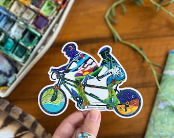 May include: A colorful sticker of a tandem bicycle with two riders. The riders are silhouetted against a landscape of mountains, trees, and stars. The bicycle has a blue and green design with flowers and leaves. The sticker has the text "@abstract.hikes" on the bottom.