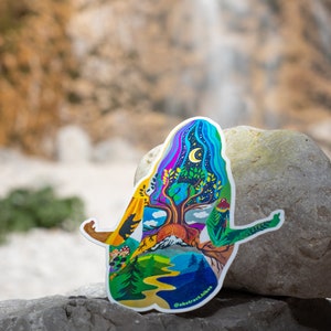 Puede incluir: Una pegatina colorida que representa una figura humana estilizada con un &aacute;rbol que crece de su pecho. La figura est&aacute; rodeada de escenas de la naturaleza, incluyendo monta&ntilde;as, un r&iacute;o y un bosque. La pegatina est&aacute; en una roca gris con una cascada borrosa en el fondo. El texto "@abstract.hikes" est&aacute; en la parte inferior de la pegatina.